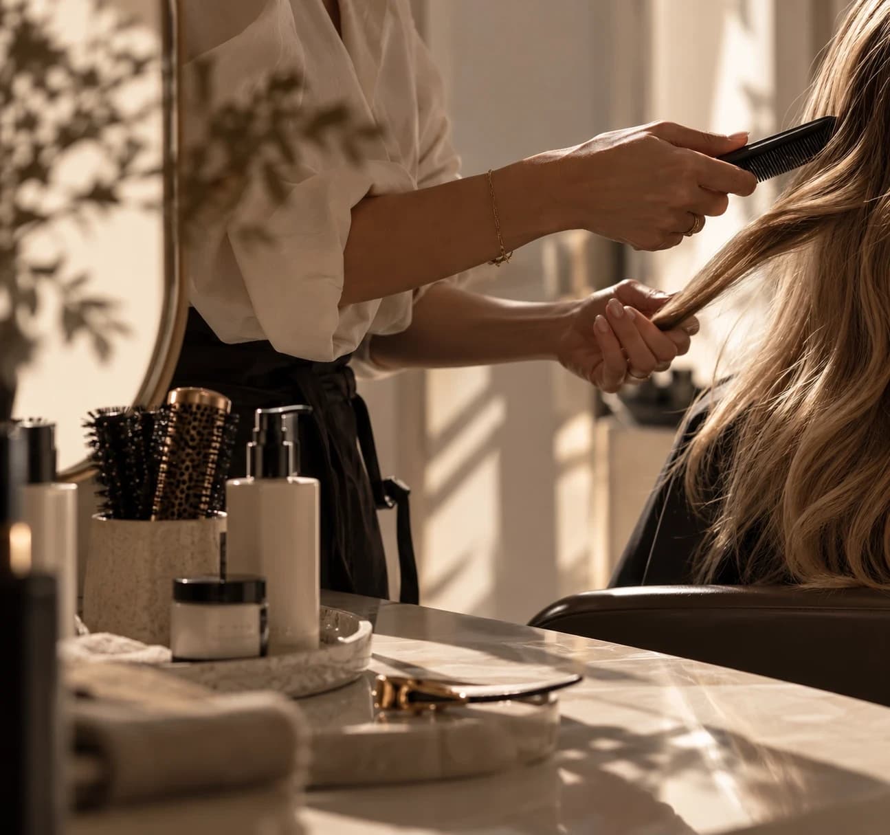 A stylist combing a client's long blonde hair beside a counter of brushes and hair-care products.
